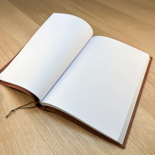 Open flexible chocolate vegetable-tanned leather journal with blank white artist-grade pages and visible hand-stitched waxed thread binding.