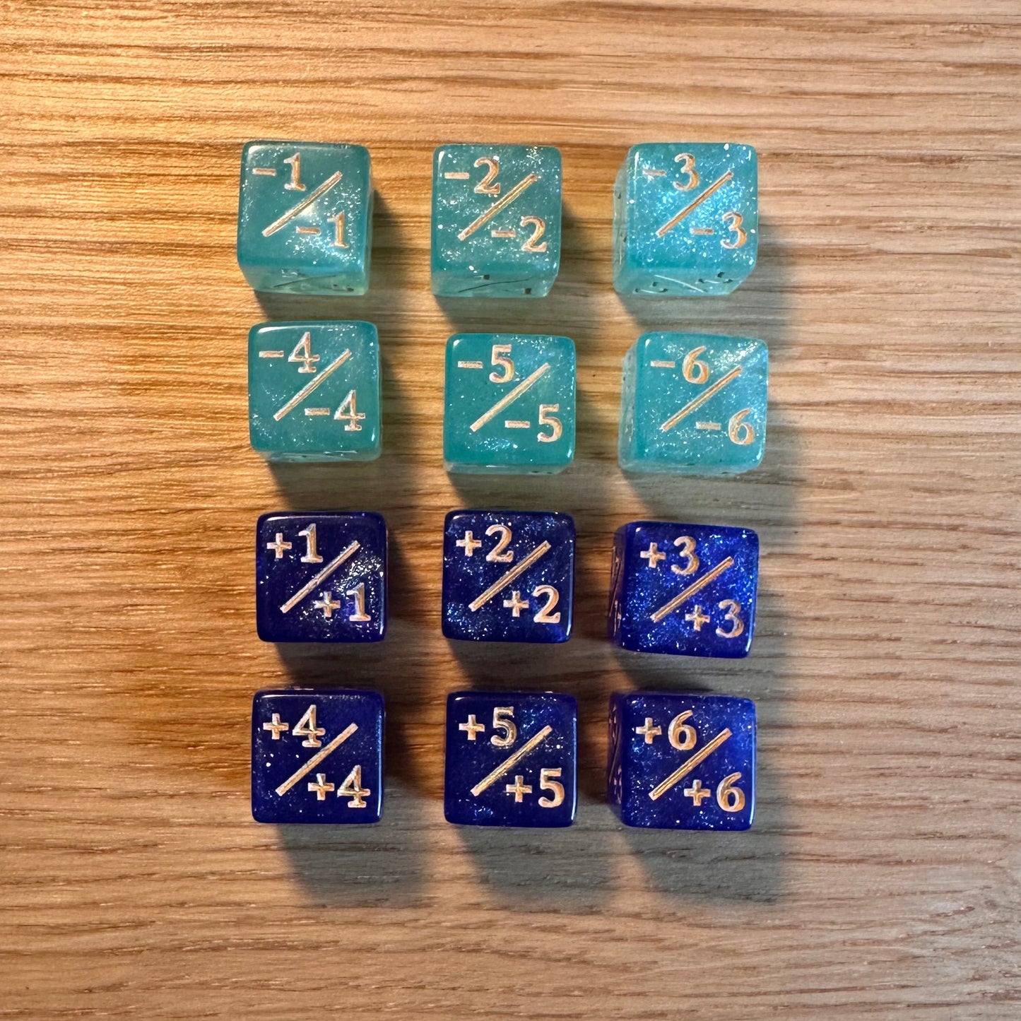 Set of 12, 12mm six-sided counter dice, including six glitter blue +/+ with gold infill dice and six glitter aqua -/- with gold infill dice, used for tracking game modifiers.