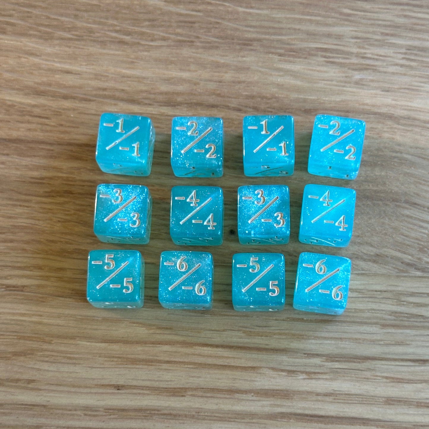 Set of twelve 12mm D6 -/- minus minus dice in glitter aqua blue with gold infill. each dice is numbered 1-6 on each side to track incremental negative modifiers.