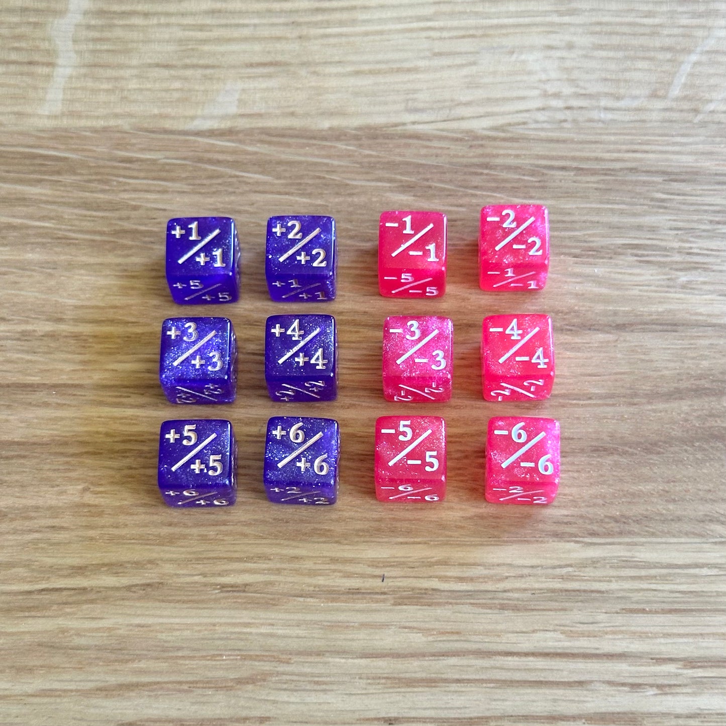 Set of 12, 12mm six-sided counter dice, including six purple glitter +/+ dice with white infill and six pink glitter -/- dice with white infill, used for tracking game modifiers.